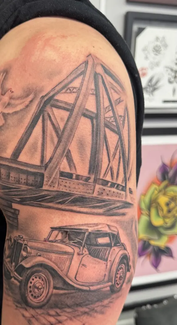 Black-and-grey tattoo on an upper arm: a steel truss bridge above a vintage convertible car on cobblestones.