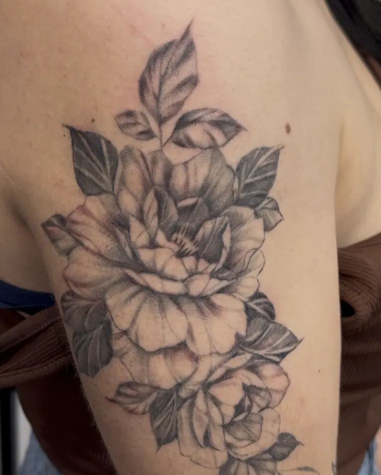 Black-and-grey peony flowers and leaves tattooed on an upper arm and shoulder, with small butterflies near the top edge.