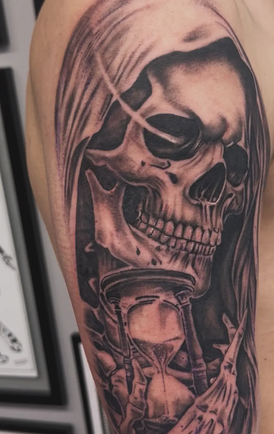 Black-and-grey tattoo on an upper arm of a hooded skull holding an hourglass.