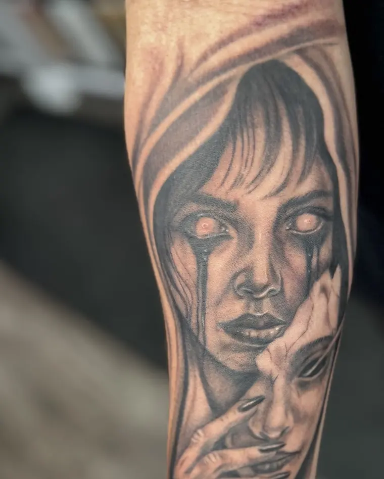 Black-and-grey tattoo on an inner forearm of a long-haired woman with tears on her face, holding a smaller mask-like face against her cheek.